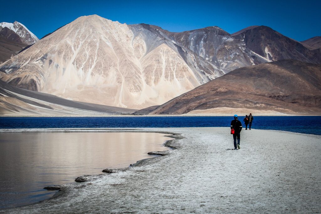 A traveler strolls along the quiet, sandy edge of Pangong Lake in Ladakh, with towering brown mountains all around and the deep blue water glistening under a bright, cloudless sky at Leh=Ladakh Trip.