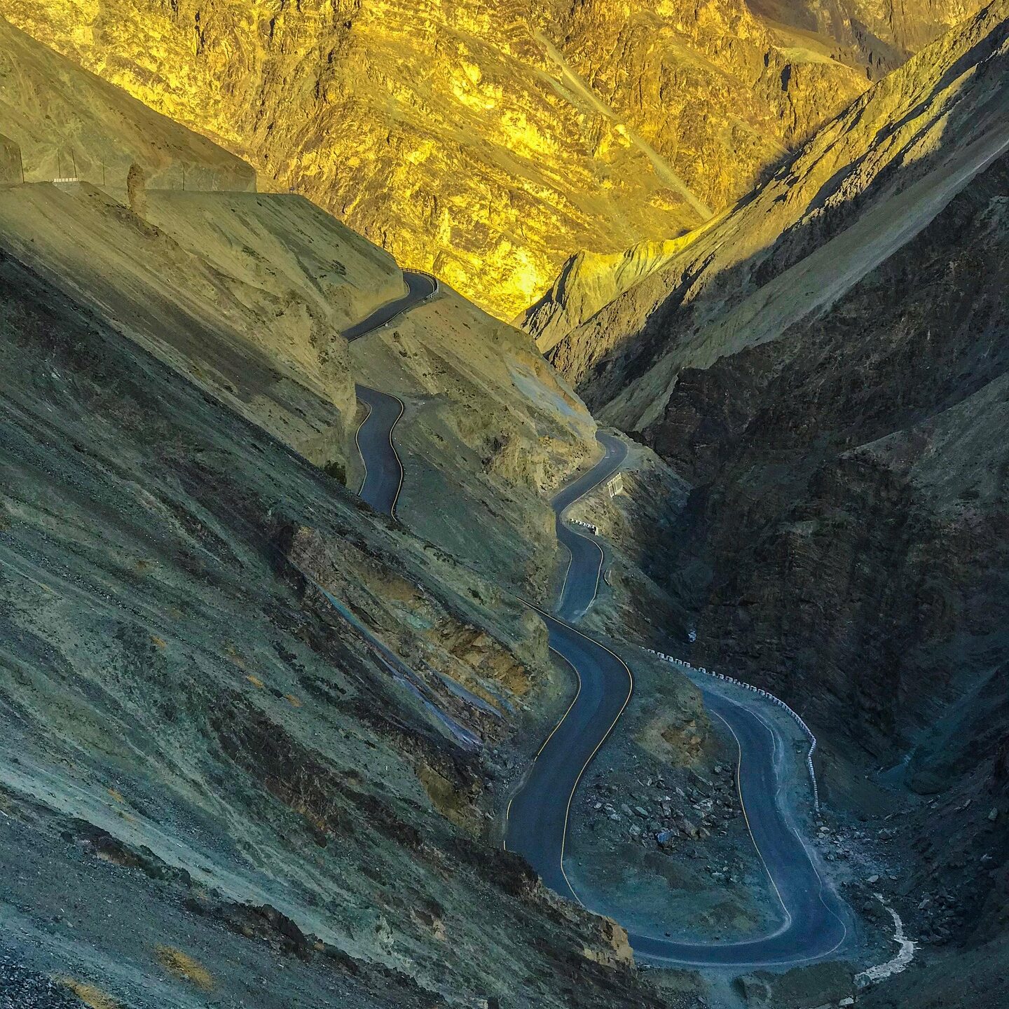 Winding mountain road cutting through rugged Ladakh valley with golden sunlight on rocky cliffs
