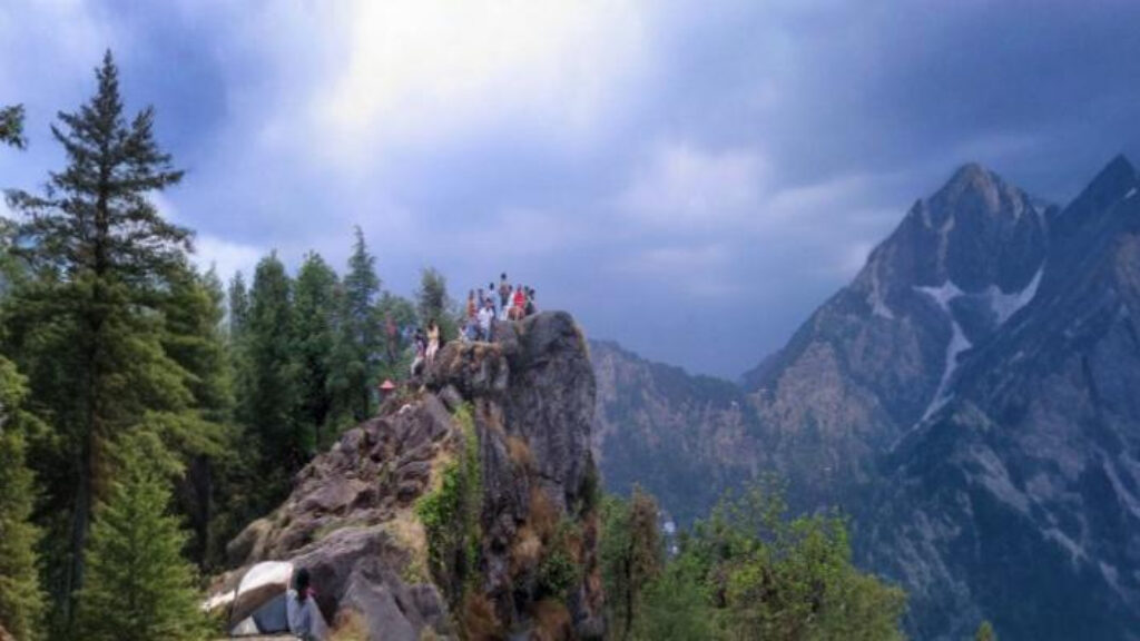 Tourists standing on Tiffin Top viewpoint surrounded by lush forest and Himalayan mountains in Nainital, India.