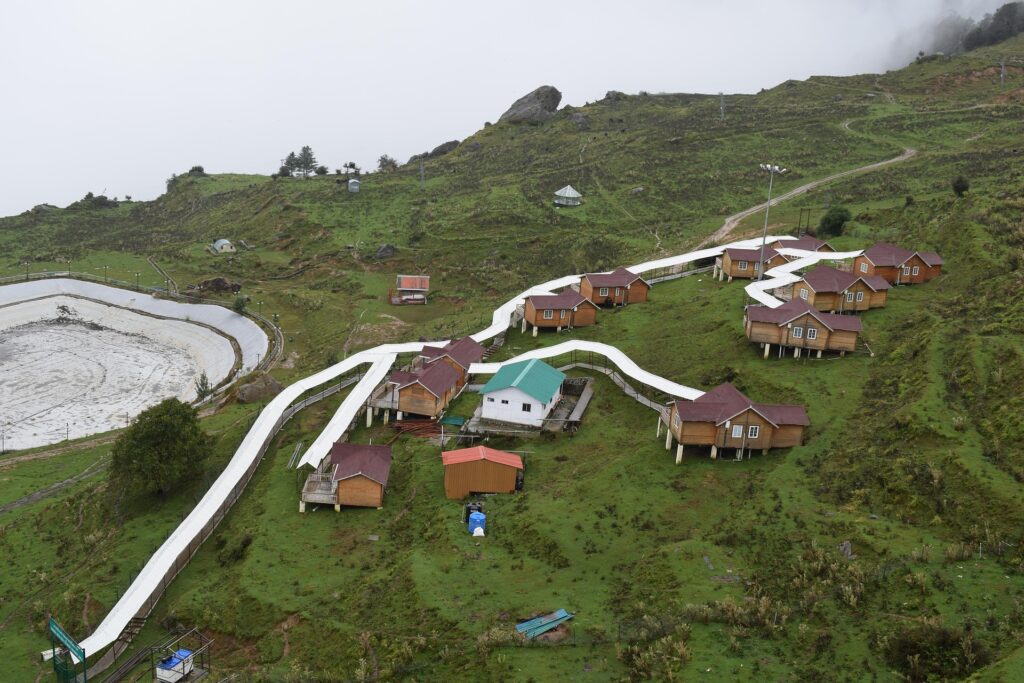 Monsoon Magic in Auli – Misty Hills, Green Slopes & Cozy Wooden Cottages Wooden cottages in Auli nestled on green hills, with low-hanging clouds and fresh monsoon mist creating a calm, peaceful vibe.